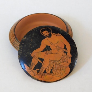 May include: A terracotta-colored ceramic bowl with a lid. The lid features a black background with an orange illustration of a seated figure holding a sword. The bowl's rim has a Greek key pattern.