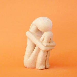 Op de afbeelding: Een beige, gladgestructureerd beeldhouwwerk van een figuur in een zittende, gekrulde houding met het hoofd gebogen naar de knieën. De vorm is abstract en afgerond, bestaande uit een massief, lichtgekleurd materiaal met een matte afwerking zonder gezicht.