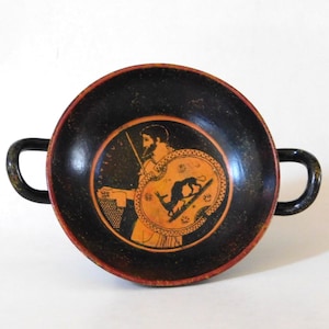 Może przedstawiać: Biały ceramiczny kubek z czarnym uchwytem i napisem "But First, Coffee" pogrubioną czcionką. Kubek jest wypełniony ciemnym płynem, prawdopodobnie kawą, i stoi na jasnej powierzchni.