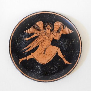 Peut inclure: Une assiette ronde en céramique noire avec une illustration détaillée d'une figure ailée en orange et blanc. La figure est représentée en pleine course, les bras tendus et des ailes détaillées. Le bord de l'assiette est orange foncé.