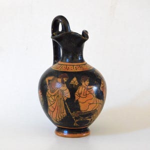 Może przedstawiać: Czarny i pomarańczowy ceramiczny dzban z uchwytem, przedstawiający starożytne greckie postacie i geometryczne wzory. Dzban ma wąską szyję i szeroką podstawę, o pojemności około 1 litra.