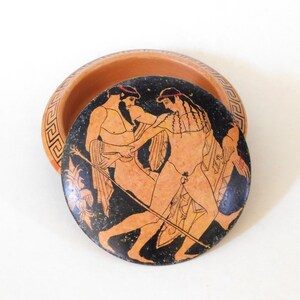 Peut inclure: Un bol en céramique de couleur terre cuite avec un couvercle orné d'une illustration noire et orange de deux personnages. Le bord du bol présente un motif de clé grecque. Le couvercle représente une scène de style classique.