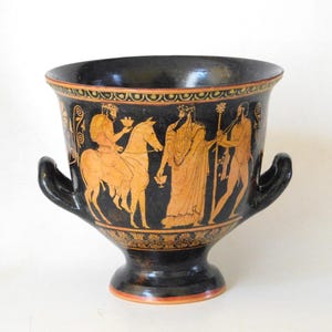 Puede incluir: Jarrón de cerámica negra con dos asas y base de pedestal. El exterior presenta figuras naranjas que representan una escena con un caballo y figuras con vestimenta clásica. Bandas decorativas en naranja y negro adornan el borde y la base.
