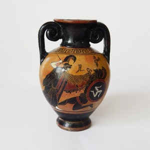 Può includere: Una piccola anfora in ceramica nera e marrone con due manici. L'anfora è decorata con una scena in bianco e nero di due figure in combattimento, una con uno scudo e l'altra con una lancia. L'anfora ha un motivo geometrico attorno al collo.