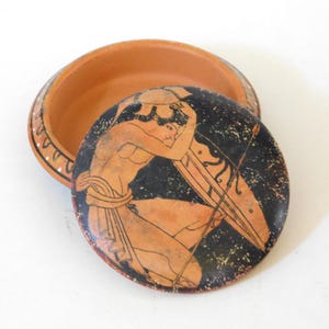 May include: A terracotta trinket box with a lid featuring a classical Greek illustration. The lid depicts a warrior with a shield and spear against a black background. The box is open, revealing its interior.