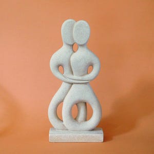 Op de afbeelding: Een lichtgekleurde abstracte sculptuur die twee omarmende figuren uitbeeldt. De figuren zijn gestileerd met gladde, afgeronde vormen en zijn verbonden bij de armen en onderlichamen. De sculptuur is gemonteerd op een rechthoekige basis.
