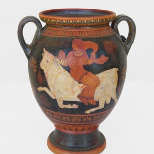 May include: A black and white ceramic vase with a red and white painted design of a woman riding a bull. The vase has two handles and a round base.