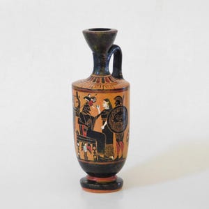 May include: A small, antique Greek vase with a black base and rim, featuring a detailed orange and black painted scene of figures. The vase has a handle and a flared top, showcasing ancient artistry.