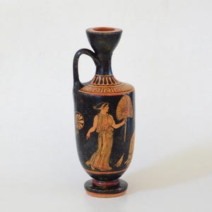 Pode incluir: Vaso de cerâmica preto e laranja com uma alça e uma parte superior alargada. O vaso apresenta um desenho pintado de uma mulher segurando um leque, com padrões de chaves gregas e outros elementos decorativos. O vaso tem aproximadamente 20 cm de altura.