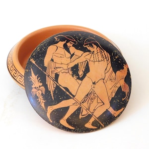 May include: A round terracotta lidded box featuring a black-figure style illustration of two figures in profile. The lid depicts a bearded individual and a youth with a staff and bird. The base has a geometric Greek key pattern along the rim.