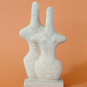 Op de afbeelding: Een lichtgekleurde stenen sculptuur van twee gestileerde figuren, dicht bij elkaar staand. De figuren hebben langwerpige halzen en afgeronde lichamen, met eenvoudige geëtste details. De sculptuur is gemonteerd op een rechthoekige basis.