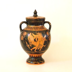 May include: A black and orange ceramic vase with a lid, featuring a classical Greek scene. The vase has two handles and a pedestal base. The artwork depicts figures in orange against a black background. The lid has a pointed finial.