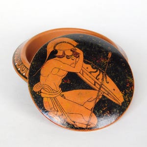 May include: A terracotta ceramic trinket box with a black and orange painted design of a Greek figure holding a spear. The lid of the box is open.