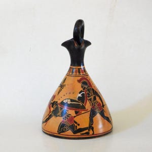 May include: A terracotta-colored Greek vase with a black handle and neck. The vase features a painted scene of warriors in black, red, and white, with the word "HEKTAP" visible. The vase has a rounded body and a flared neck.