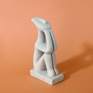 Op de afbeelding: Een lichtgrijze abstracte sculptuur van een zittende figuur met de armen om de knieën. De sculptuur staat op een rechthoekige basis. Het hoofd van de figuur is iets omhoog gekanteld. De achtergrond is effen oranje.