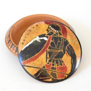May include: A terracotta-colored trinket box with a lid featuring a painted warrior scene. The warrior is depicted in black and red, holding a shield and sword against a yellow background. The lid is inscribed with the word "AXIVEVS".
