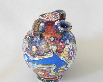 Pode incluir: Um vaso de cerâmica pintado à mão com corpo redondo, alça e bico. O vaso apresenta um desenho de golfinho azul, com detalhes em vermelho, azul e amarelo. O vaso tem aproximadamente 20 cm de altura.