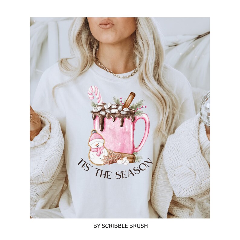 May include: White sweatshirt with a pink and brown watercolor illustration of a mug of hot chocolate with marshmallows, a snowman, and cinnamon sticks. The text "Tis' the Season" is printed below the mug. The text "By Scribble Brush" is printed at the bottom of the sweatshirt.