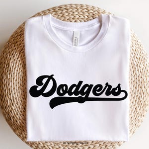K&ouml;nnte beinhalten: Wei&szlig;es T-Shirt mit dem Wort "Dodgers" in schwarzer, retro Schrift. Das T-Shirt liegt auf einer gewebten, kreisf&ouml;rmigen Oberfl&auml;che. Der Text "by scribble brush" befindet sich unten. Das T-Shirt ist aus weichem Material.