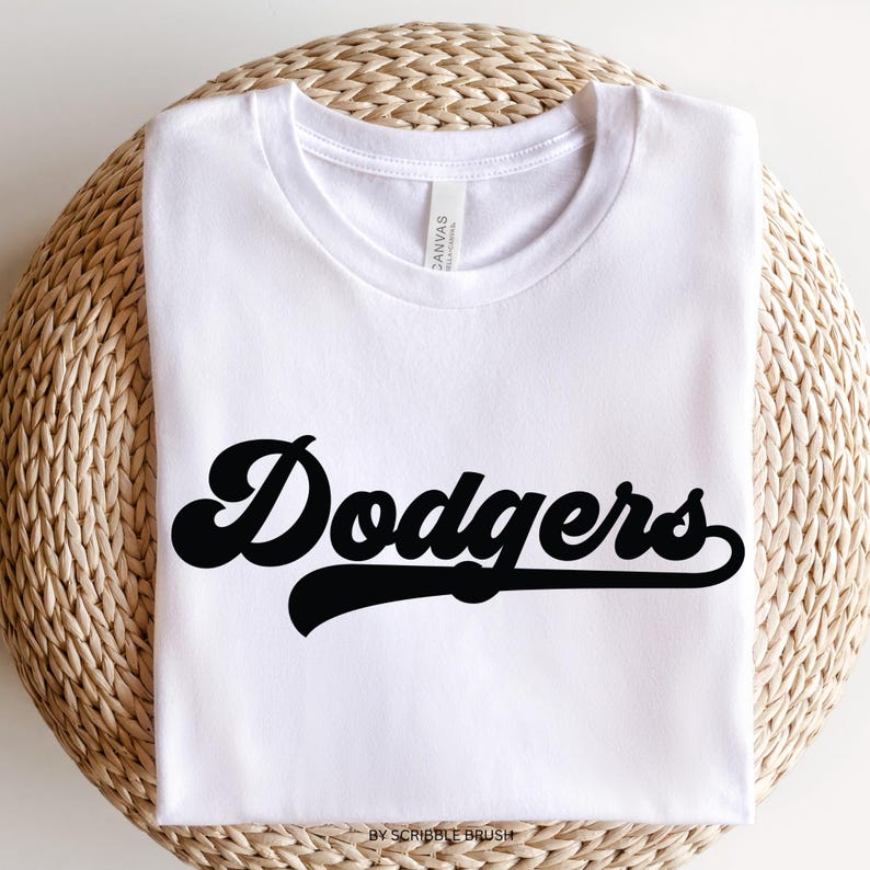 K&ouml;nnte beinhalten: Wei&szlig;es T-Shirt mit dem Wort "Dodgers" in schwarzer, retro Schrift. Das T-Shirt liegt auf einer gewebten, kreisf&ouml;rmigen Oberfl&auml;che. Der Text "by scribble brush" befindet sich unten. Das T-Shirt ist aus weichem Material.