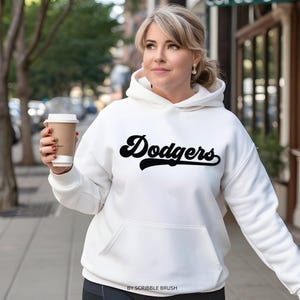 K&ouml;nnte beinhalten: Ein wei&szlig;er Kapuzenpullover mit dem Wort "Dodgers" in schwarzer Schrift auf der Vorderseite. Der Kapuzenpullover hat eine Vordertasche und eine Kapuze mit Kordelzug. Das Bild enth&auml;lt auch den Text "BY SCRIBBLE BRUSH" unten.