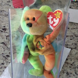 May include: A multi-coloured tie-dye beanie baby bear with a peace sign on its chest. The bear is in a clear plastic display case with a red Ty beanie baby tag that says "Beanie Babies Baby" and has the Ty logo.