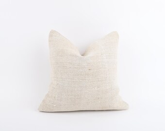 May include: A square, off-white pillow with a textured, woven appearance. The pillow is centered on a white background, showcasing its neutral color and simple design. The pillow's shape is symmetrical, with a soft, inviting look.