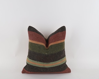 May include: A square pillow with a striped pattern in shades of brown, red, green, and orange. The pillow has a textured appearance and is likely made of woven fabric. The stripes run horizontally across the pillow.