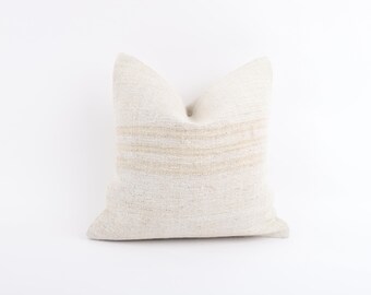 May include: A square, off-white decorative pillow with horizontal tan stripes. The pillow has a textured appearance and a soft, neutral color palette, suitable for various interior design styles. The pillow is approximately 18 inches square.