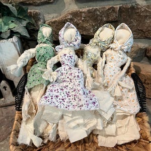 May include: A collection of handmade fabric dolls in a woven basket. The dolls have fabric dresses and bonnets in various floral patterns and colors, including green, purple, and yellow. The dolls' arms are made of twisted fabric.