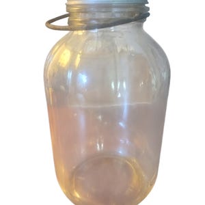 Op de afbeelding: Een heldere glazen pot met een zilveren metalen deksel en een draadhandvat. De pot heeft een afgeronde body en een smalle hals. Het deksel is bevestigd met een metalen ring. De pot is leeg en lijkt vintage.