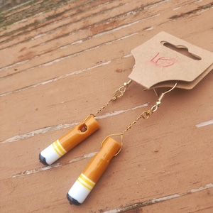 Op de afbeelding: Een paar novelty oorbellen die ontworpen zijn om op sigaretten te lijken. Elke oorbel heeft een bruine buis met een witte punt, gele strepen en een zwart uiteinde. De oorbellen hangen aan goudkleurige oorhaken en kettingen.