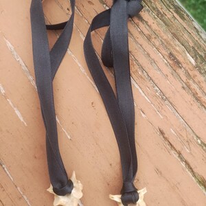 Op de afbeelding: Twee kettingen met benen hangers en zwarte linten. De hangers zijn lichtbeige van kleur en de linten zijn effen zwart. De kettingen worden gepresenteerd op een verweerd houten oppervlak.