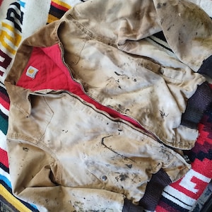 May include: A worn, tan work jacket with a red quilted lining and a brown corduroy collar. The jacket has a zipper closure and ribbed cuffs. The jacket is heavily stained with dark spots.