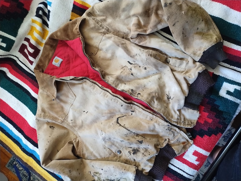 May include: A worn, tan work jacket with a red quilted lining and a brown corduroy collar. The jacket has a zipper closure and ribbed cuffs. The jacket is heavily stained with dark spots.