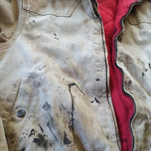 May include: A well-worn, tan work jacket with visible dark stains and a brown corduroy collar. The jacket features a full zipper and a red quilted lining. A metal button secures the front pocket.