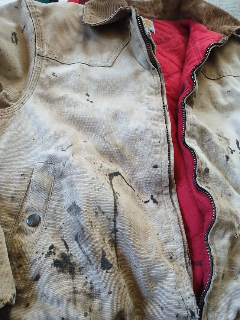May include: A well-worn, tan work jacket with visible dark stains and a brown corduroy collar. The jacket features a full zipper and a red quilted lining. A metal button secures the front pocket.