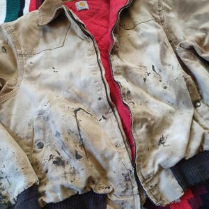 May include: A tan work jacket with a brown corduroy collar and a red quilted lining. The jacket has a zipper closure and shows signs of wear with dark stains. The cuffs and waistband are a dark brown ribbed knit.