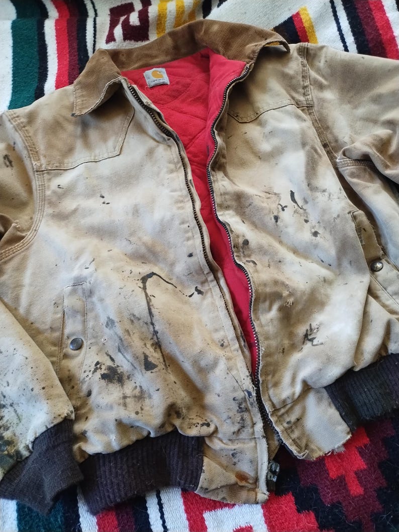 May include: A tan work jacket with a brown corduroy collar and a red quilted lining. The jacket has a zipper closure and shows signs of wear with dark stains. The cuffs and waistband are a dark brown ribbed knit.