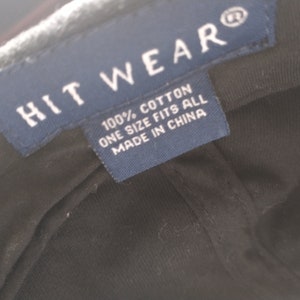 Puede incluir: Primer plano de una gorra de b&eacute;isbol negra con una etiqueta azul. La etiqueta dice "HIT WEAR" y "100% ALGOD&Oacute;N TALLA &Uacute;NICA FABRICADO EN CHINA". La gorra es de algod&oacute;n.