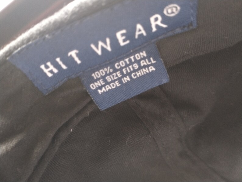 Puede incluir: Primer plano de una gorra de b&eacute;isbol negra con una etiqueta azul. La etiqueta dice "HIT WEAR" y "100% ALGOD&Oacute;N TALLA &Uacute;NICA FABRICADO EN CHINA". La gorra es de algod&oacute;n.