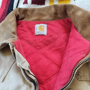 May include: Close-up of a Carhartt work jacket. The jacket is tan with a brown corduroy collar and a red quilted lining. The Carhartt logo is visible on a tag inside the jacket. A metal zipper runs down the front.