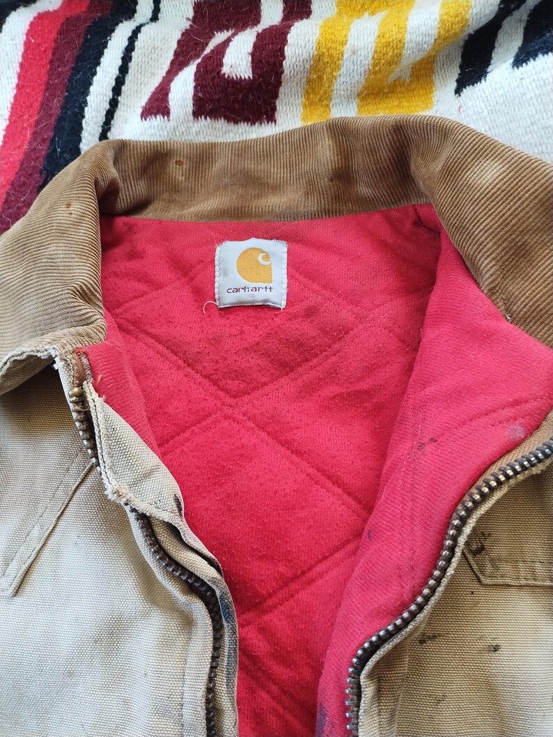 May include: Close-up of a Carhartt work jacket. The jacket is tan with a brown corduroy collar and a red quilted lining. The Carhartt logo is visible on a tag inside the jacket. A metal zipper runs down the front.