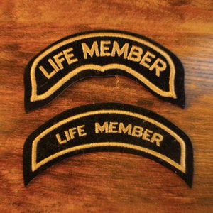 Könnte beinhalten: Zwei schwarze, gestickte Aufnäher mit dem Text "LIFE MEMBER" in goldfarbenen Buchstaben. Die Aufnäher haben eine gebogene Form und einen goldenen Rand. Die Aufnäher werden auf einer Holzoberfläche präsentiert.