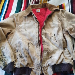 May include: A tan work jacket with a brown corduroy collar and a red quilted lining. The jacket has a zipper closure, two front pockets, and dark brown ribbed cuffs. The jacket shows signs of wear with dark stains and a Carhartt logo.