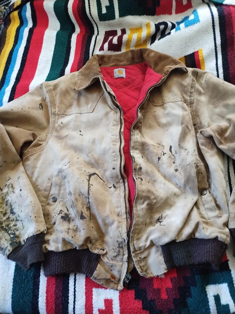 May include: A tan work jacket with a brown corduroy collar and a red quilted lining. The jacket has a zipper closure, two front pockets, and dark brown ribbed cuffs. The jacket shows signs of wear with dark stains and a Carhartt logo.