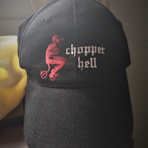 Puede incluir: Gorra de béisbol negra con visera curva. La gorra presenta un diablo rojo bordado y las palabras "chopper hell" en letras góticas blancas. La gorra está hecha de tela texturizada.