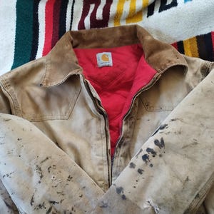 May include: A tan work jacket with a brown corduroy collar and a red quilted lining. The jacket has a zipper closure and a small logo patch. The jacket shows signs of wear with dark stains.