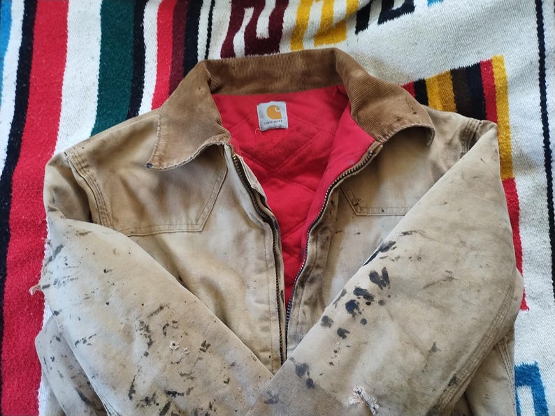 May include: A tan work jacket with a brown corduroy collar and a red quilted lining. The jacket has a zipper closure and a small logo patch. The jacket shows signs of wear with dark stains.