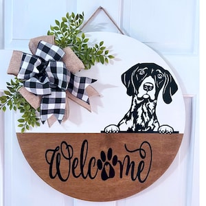 May include: A round wooden door hanger with a white top half and a brown bottom half. The top half features a black and white silhouette of a dog peeking over the edge. The bottom half has the word "Welcome" written in black with a paw print in the middle. A black and white checkered bow with a burlap bow is on the top left side of the door hanger.